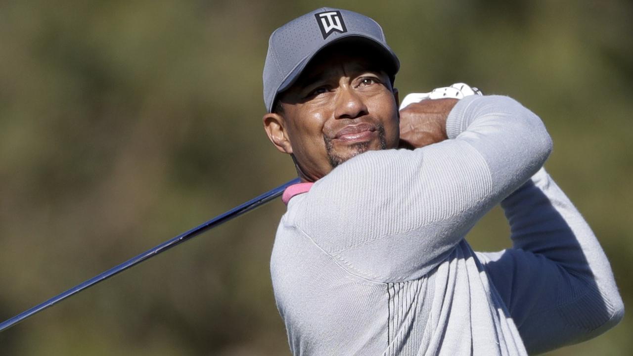 Woods hat gerade abgeschlagen und den Schläger hinter den Kopf geschwungen. Er trägt einen hellgrauen Pulli und eine dunkelgraue Baseballkappe und schaut dem Ball mit skeptischem Blick hinterher.