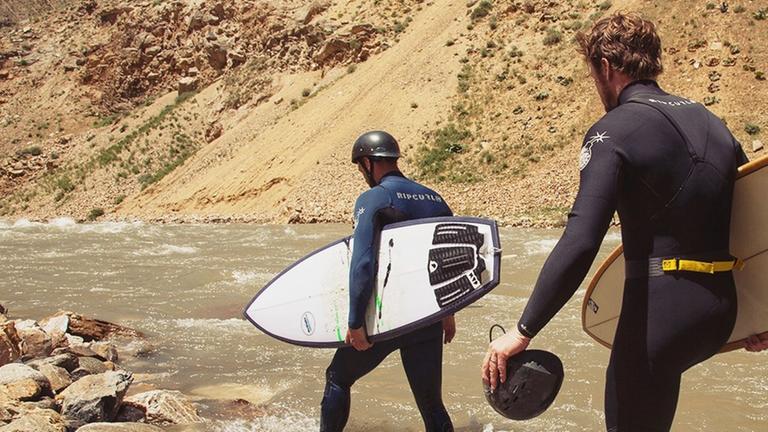 Zwei Männer in Neoprenanzügen und mit Surfboards in den Händen waten durch einen Fluss in Afghanistan.