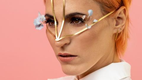 Eine Frau mit pfirsichfarbenem kurzen Haar trägt ein feines goldenes Gestell über Stirn und Nase. Hellblaue Blumen wirken wie Wimpernverlängerungen. Sie trägt eine weiße Bluse mit Kragen, steht vor rosafarbenem Hintergrund und blickt mit konzentriertem Blick seitlich an der Kamera vorbei.