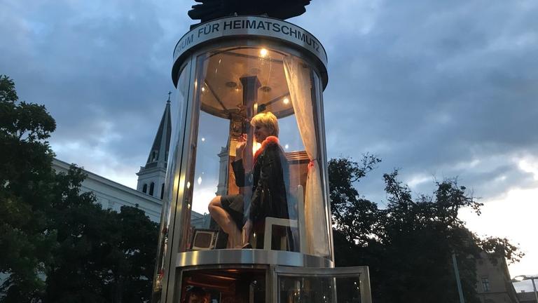 Eine Litfaßsäule aus Glas, in ihr sitze eine blonde Frau.
