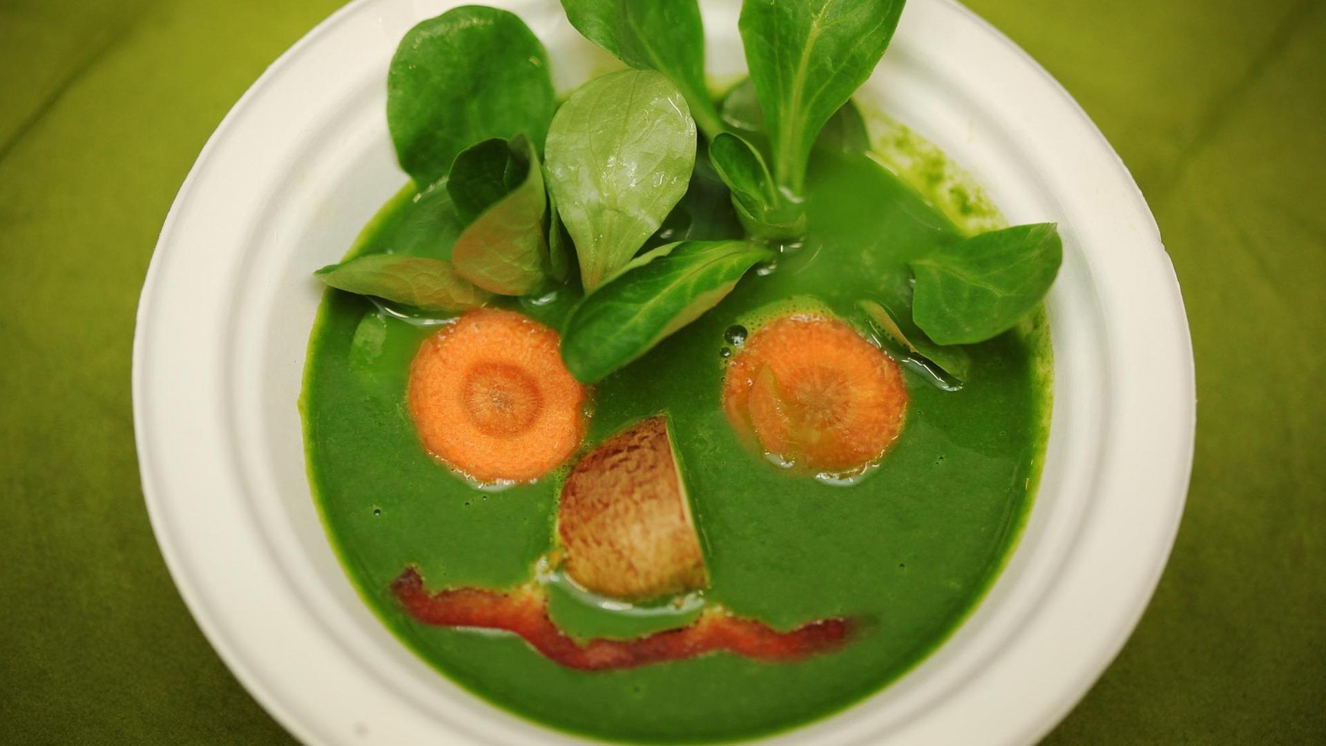 Eine vegane Spinat-Suppe, auf die aus Gemüse wie Spinatblätter und Möhren ein Gesicht gezeichnet wurde.
