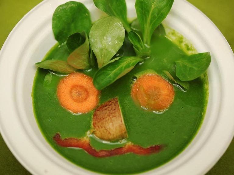 Eine vegane Spinat-Suppe, auf die aus Gemüse wie Spinatblätter und Möhren ein Gesicht gezeichnet wurde.