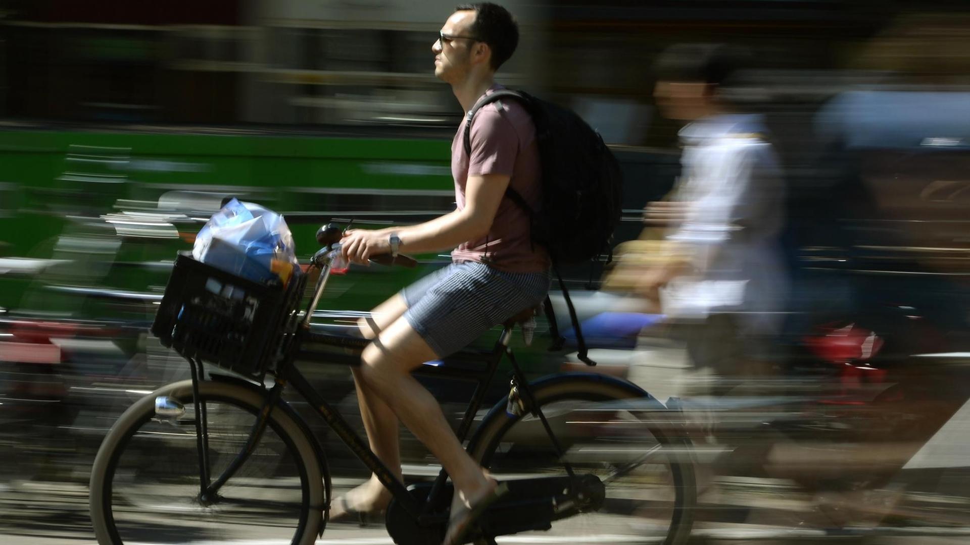 Ein Mann transport in seinem Fahrradkorb vorne am Lenker seine Einkäufe, während entlang einer der Amsterdamer Grachten fährt.