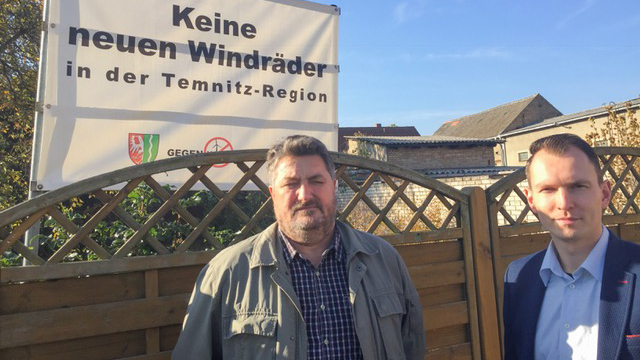 Thomas Voigt (li), der Bürgermeister von Temnitztal, und sein Stellvertreter Michael Mann (re) stehen vor einem Plakat, auf dem steht "Keine neuen Windräder in der Temnitz-Region".