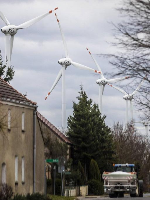 Blick auf eine Dorfstraße in Feldheim (Brandenburg) Auf der Straße fährt ein LKW, links schmucklose grau-braun gestrichene Häuser, im Hintergrund die Windräder eines Windparks.