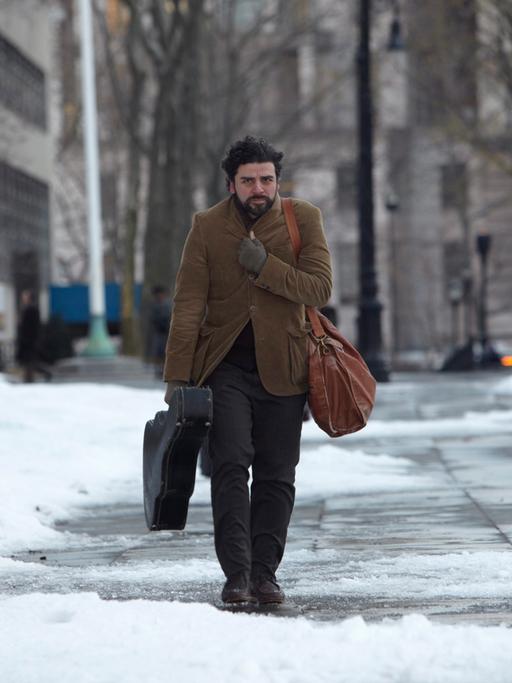 Film-Still: US-Schauspieler Oscar Isaac spaziert in "Inside Llewyn Davis" durch eine schneebedeckte Straße, Gitarre in der Hand, sich mit dem Sakko gegen die Kälte schützend