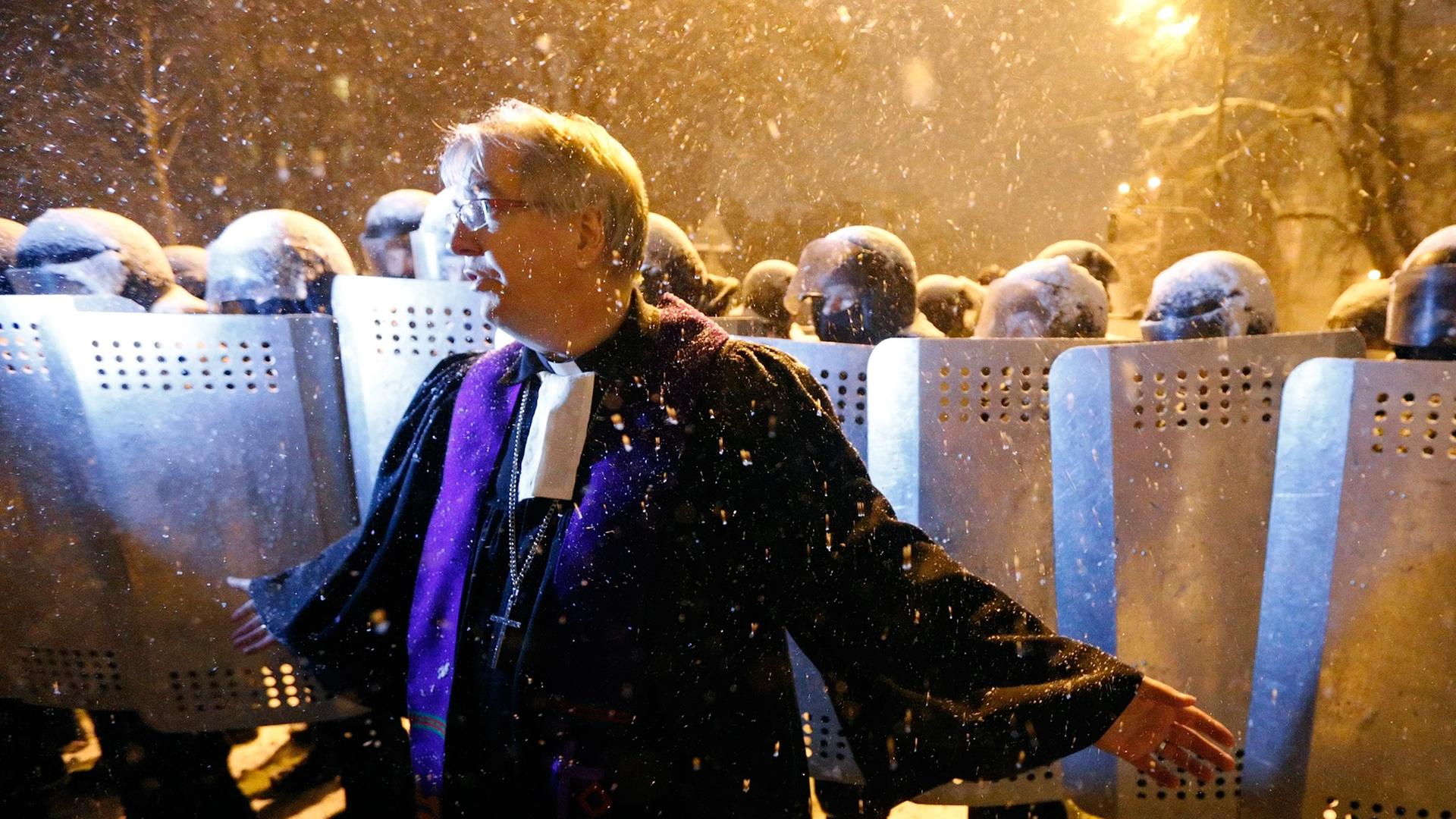 Pfarrer Ralf Haska steht in violettem Ornat vor einer Reihe von ukrainischen Sicherheitskräften, die sich hinter Schutzschilden verschanzt haben. Es ist Nacht, und es schneit.