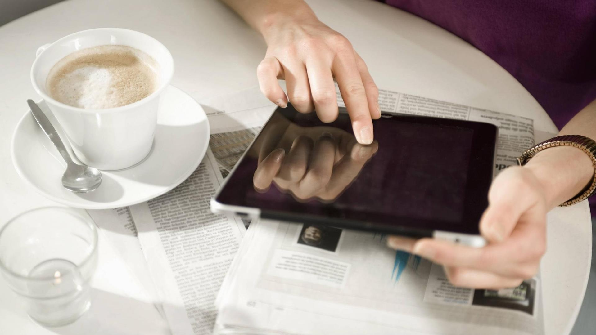 Eine Frau sitzt in einem Café und benutzt ein Tablet, das auf auf einer Zeitung liegt.