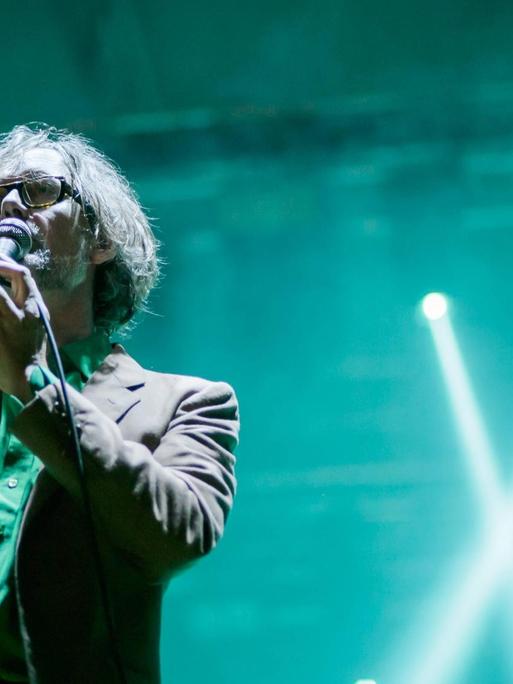 Der Sänger Jarvis Cocker steht mit Mikrofon auf einer in grün-blaues Licht getauchten Konzertbühne.