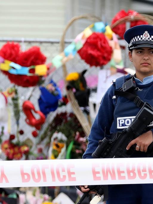 Eine Polizistin steht vor dem abgesperrten Tatort des Terroranschlags in Christchurch, hinter ihr sind zahlreiche niedergelegte Blumen zu sehen.