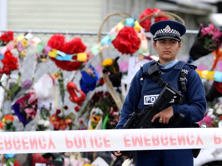Eine Polizistin steht vor dem abgesperrten Tatort des Terroranschlags in Christchurch, hinter ihr sind zahlreiche niedergelegte Blumen zu sehen.