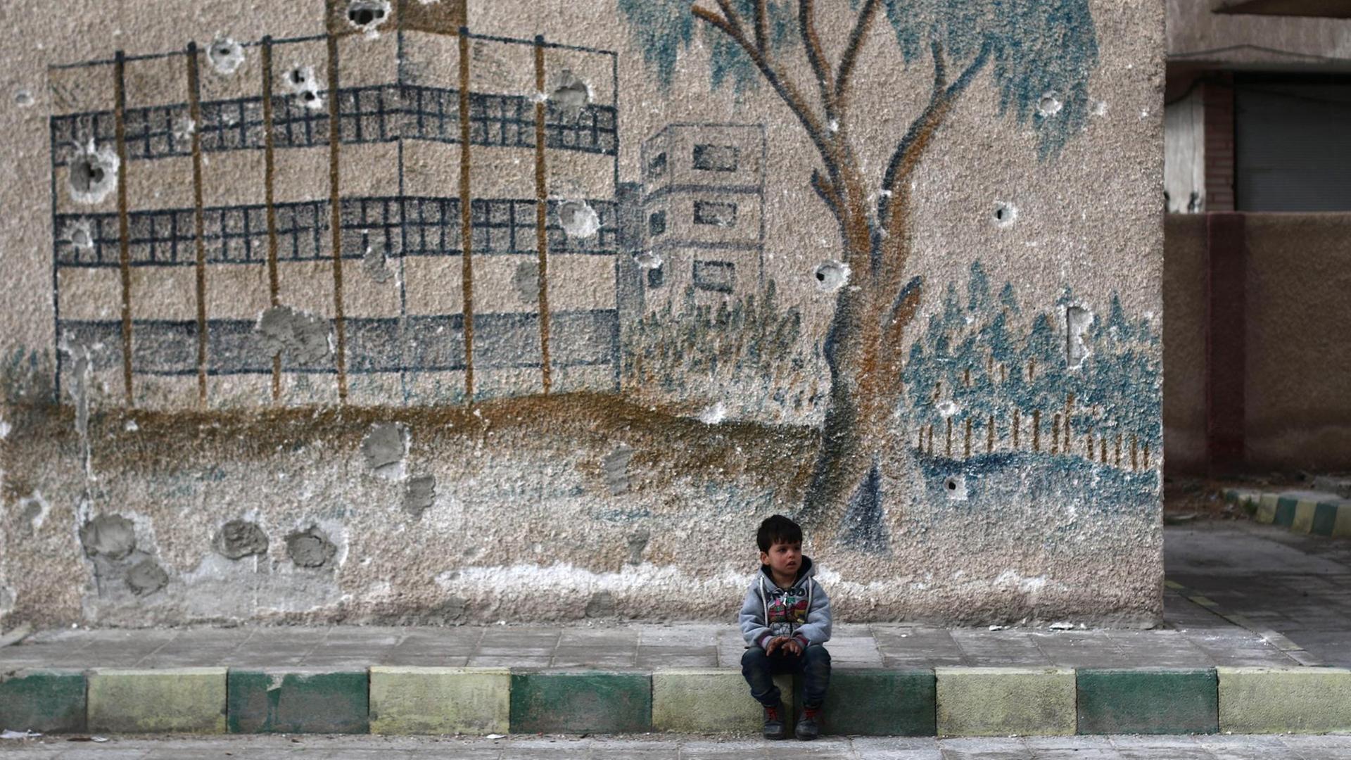 Ein kleiner Junge sitzt vor einem grauen Schulgebäude mit einer Wandmalerei auf dem Bordstein.