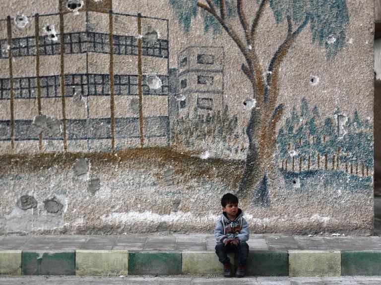 Ein kleiner Junge sitzt vor einem grauen Schulgebäude mit einer Wandmalerei auf dem Bordstein.