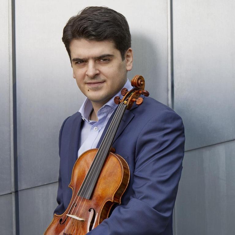 Ein Mann mit dunklen Haaren in Hemd und dunkelblauem Anzug steht vor einer grauen Wand und hält eine Geige vor seinem Körper. Es handelt sich um Michael Barenboim.