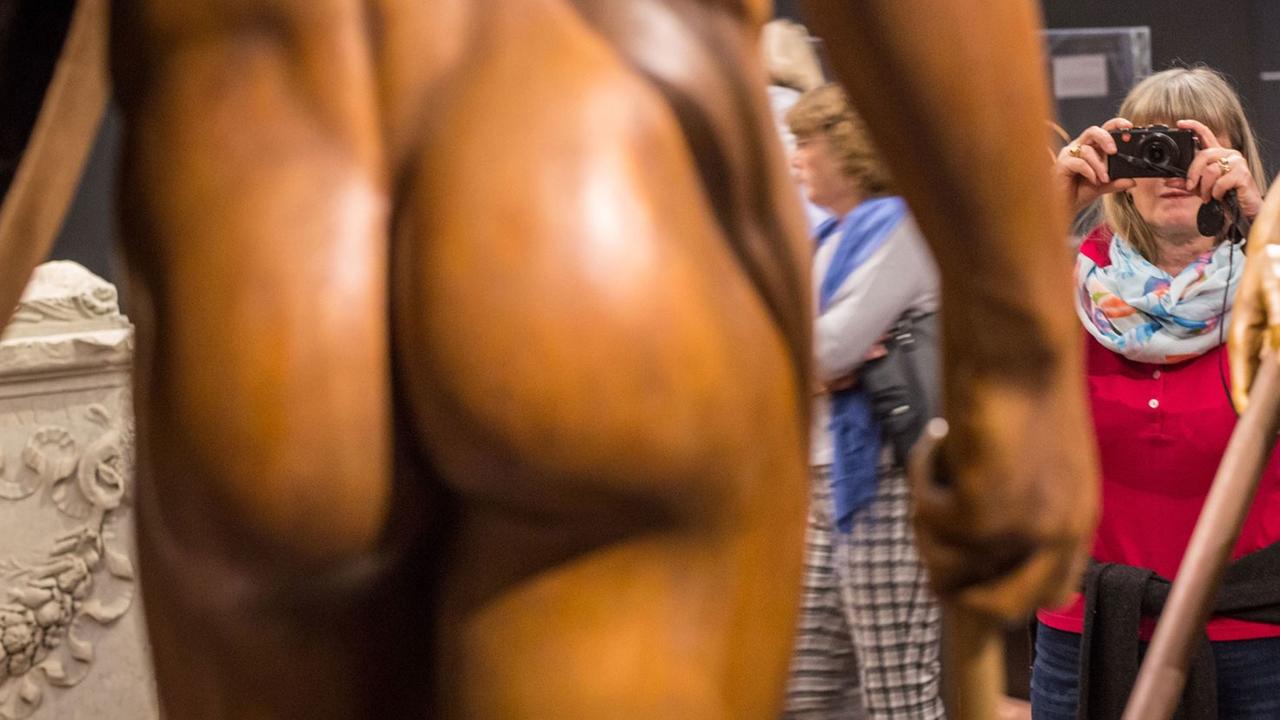 Der Po einer Männer-Skulptur in Athen ist zu sehen. Daneben eine Frau, die fotografiert.