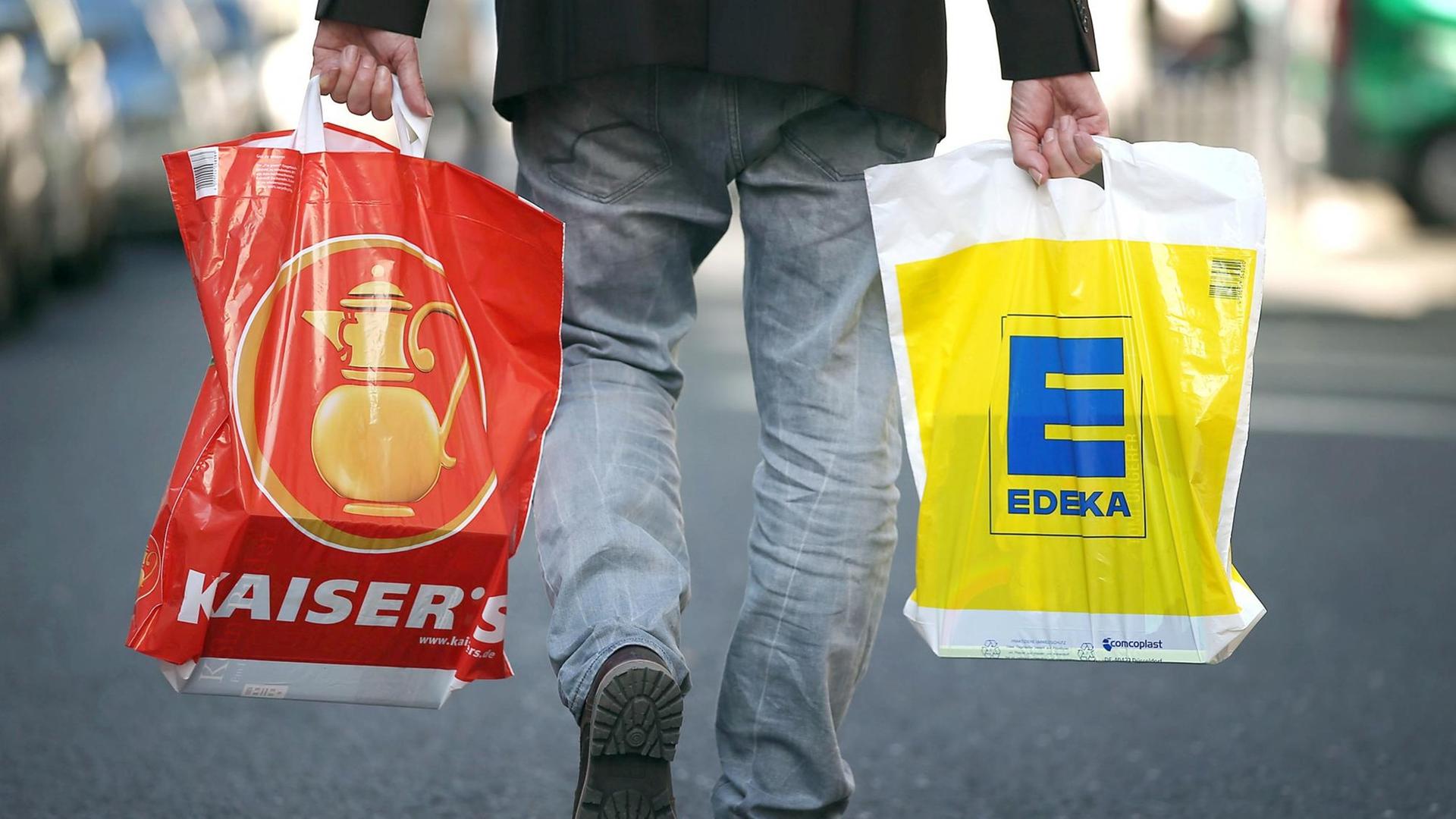 Ein Mann geht mit jeweils einer Tragetasche der Supermarktketten Kaisers's Tengelmann und Edeka über die Straße.