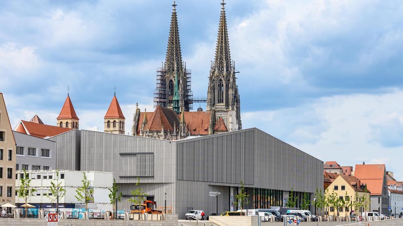 Das Museum der Bayerischen Geschichte am Regensburger Donauufer. Ein architektonisch postmodernes Gebäude am Ufer eines Flusses hebt sich ab vor einem Dom im Hintergrund.
