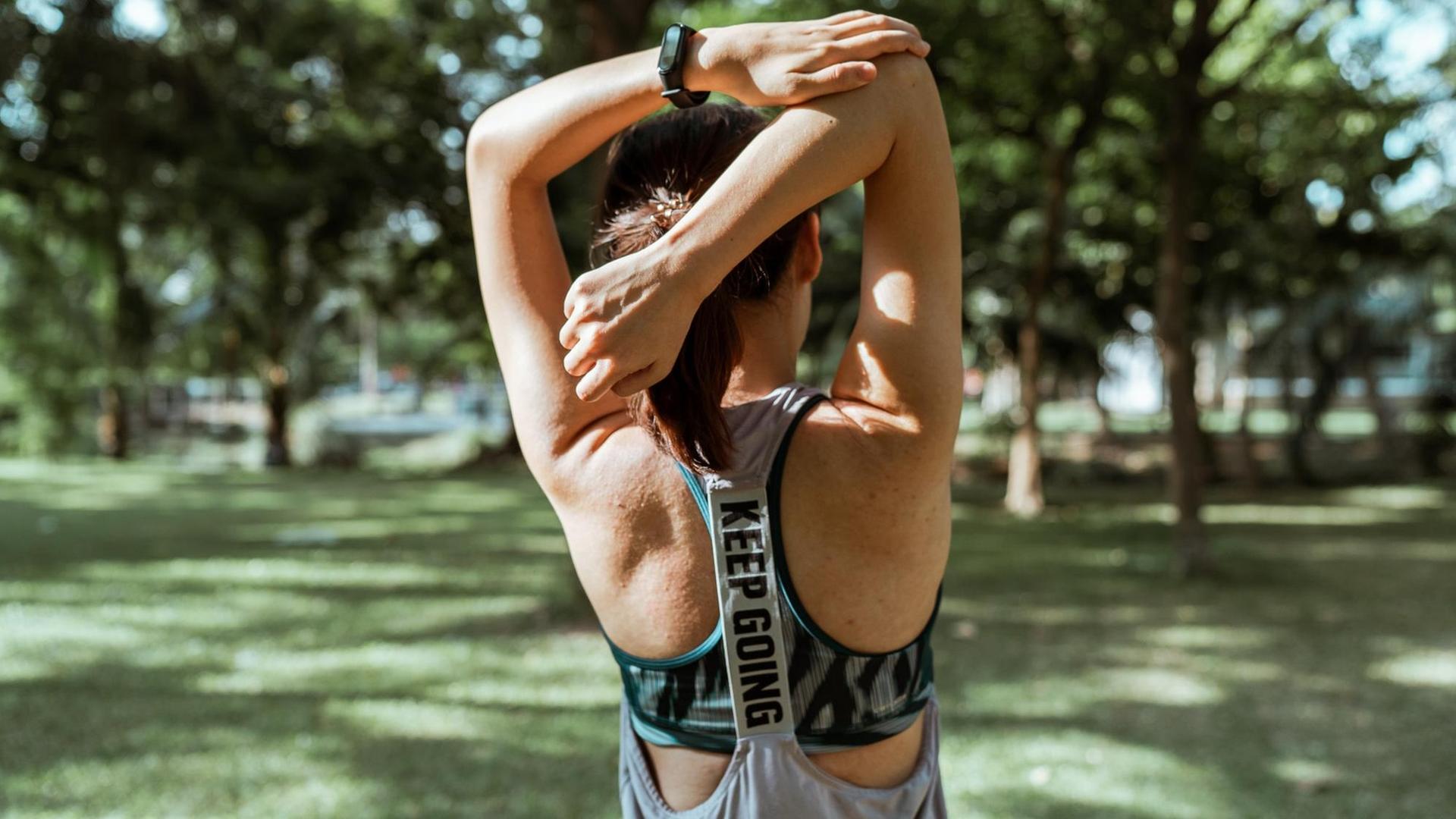 Eine Frau steht im Park und dehnt ihre Arme über den Kopf zum Rücken.