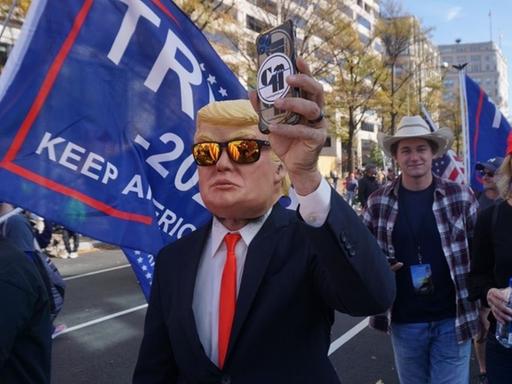 Ein Anhänger von Donald Trump trägt eine Maske Trumps und hält ein Smartphone in die Höhe