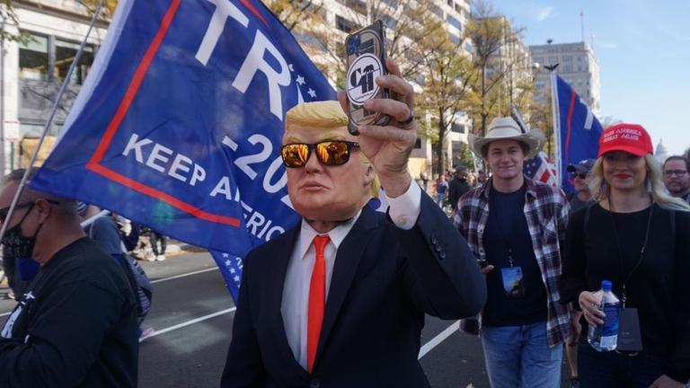 Ein Anhänger von Donald Trump trägt eine Maske Trumps und hält ein Smartphone in die Höhe