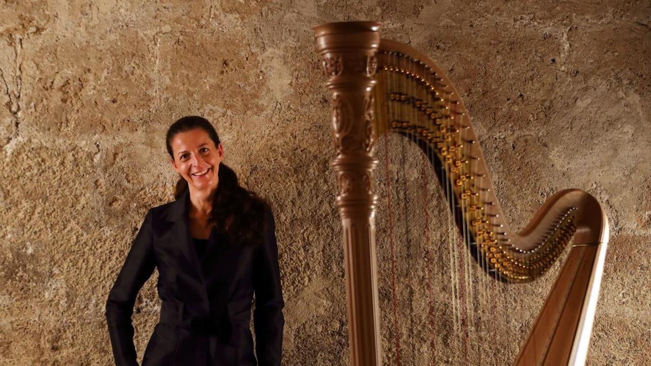 Eine junge Frau in schwarzem Hosenanzug steht etwas hinter ihrer großen Konzertharfe und lächelt.