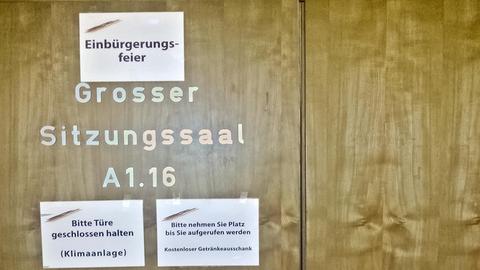 Das Bild zeigt eine verschlossene Holztür auf der Großer Sitzungssaal A1.16 steht. Desweiteren hängen drei Schilder an der Tür auf denen Einbürgerungsfeier, Bitte Türe geschlossen halten (Klimaanlage) und Bitte nehmen Sie Platz bis Sie aufgerufen werden - Kostenloser Getränkeausschank zu lesen ist.