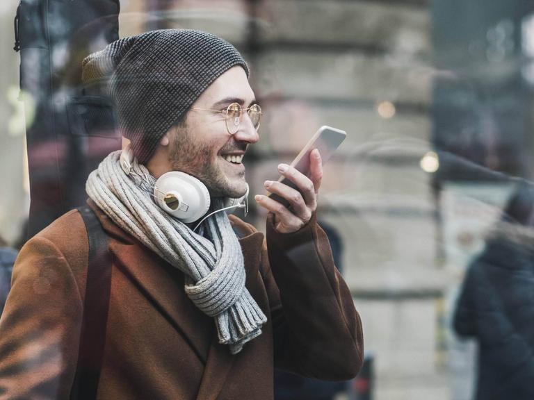 Ein lachender junger Mann mit Schal, Mütze und Gitarre auf dem Rücken spricht eine Nachricht in sein Smartphone (Symbolfoto)