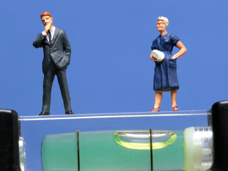 Zwei Miniatur-Figuren, eine männlich, eine weiblich, stehen auf einer schiefen Ebene einer Wasserwaage. Die männliche Figur steht etwas weiter unten.