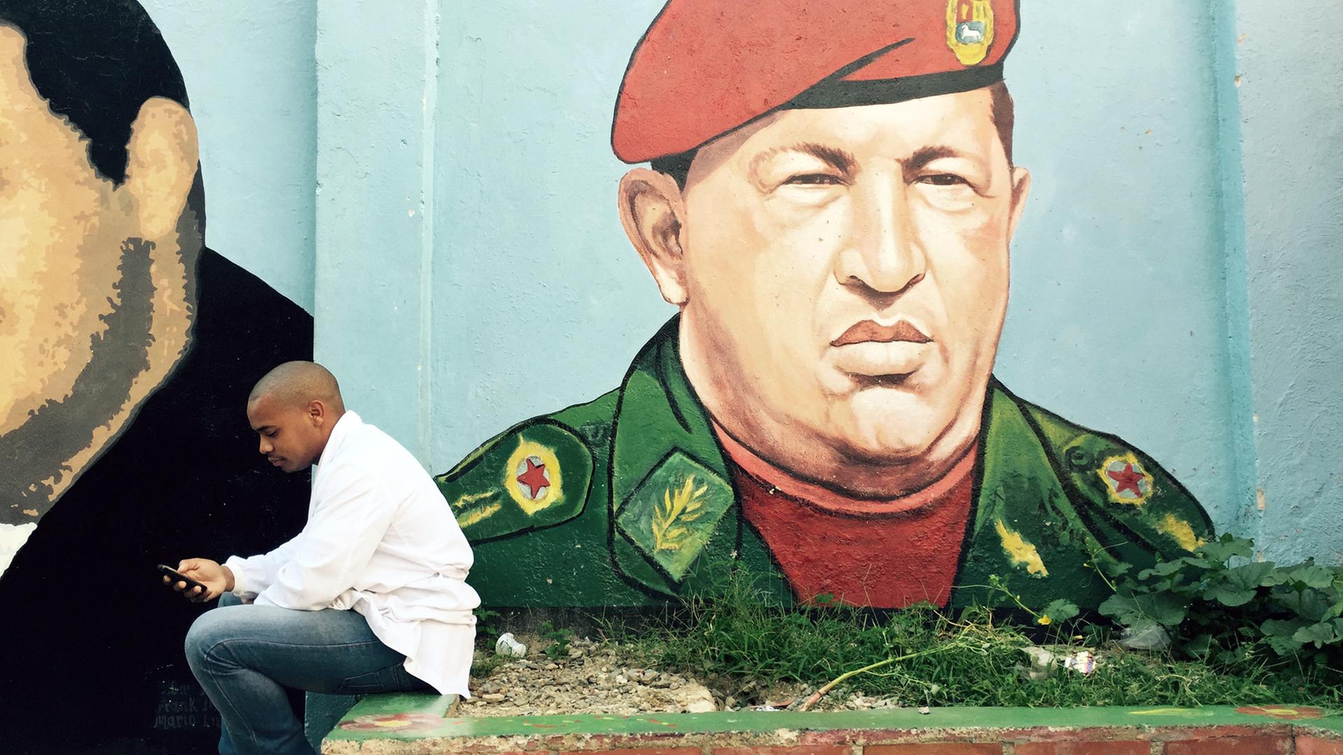 Ein junger Mann sitzt vor einem Graffiti von Hugo Chávez
