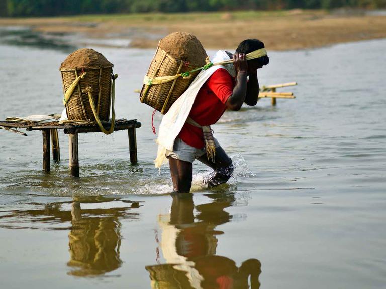 Eine Mann watet aus dem Wasser, auf dem Rücken trägt er einen Eimer voll Sand.