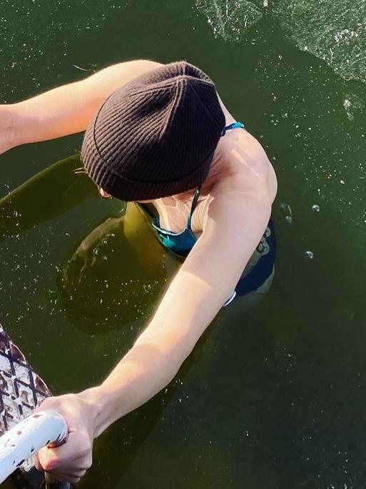 Eine Frau beim Eisbaden im See; nach dem Bad steigt sie die Leiter hoch, auf dem Wasser ist eine dünne Eisschicht zu sehen.