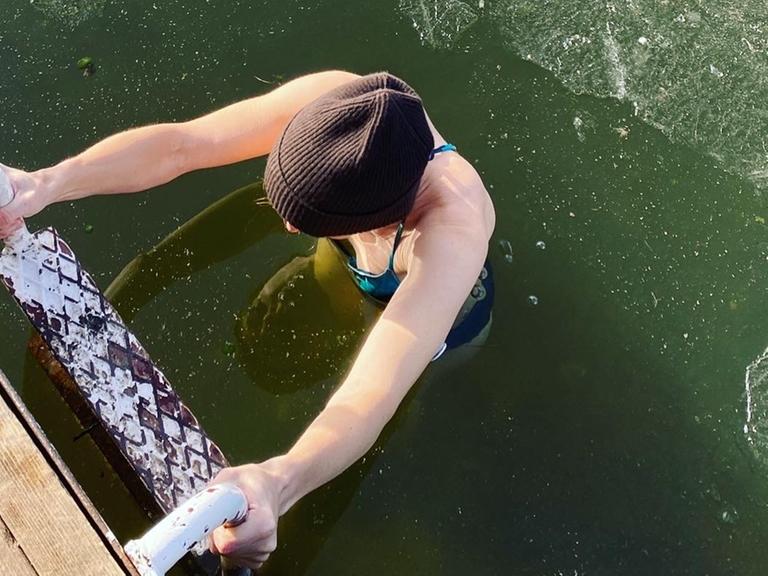 Eine Frau beim Eisbaden im See; nach dem Bad steigt sie die Leiter hoch, auf dem Wasser ist eine dünne Eisschicht zu sehen.