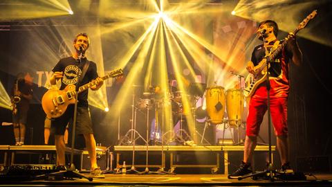 Fünf Männer stehen auf einer hell ausgeleuchteten Bühne und spielen Musik.