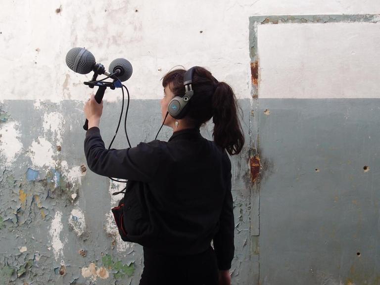 Die Klangkünstlerin Marie Guérin steht vor einer Wand im Halbmondlager in Wünsdorf und nimmt mit einem Mikrofon auf.