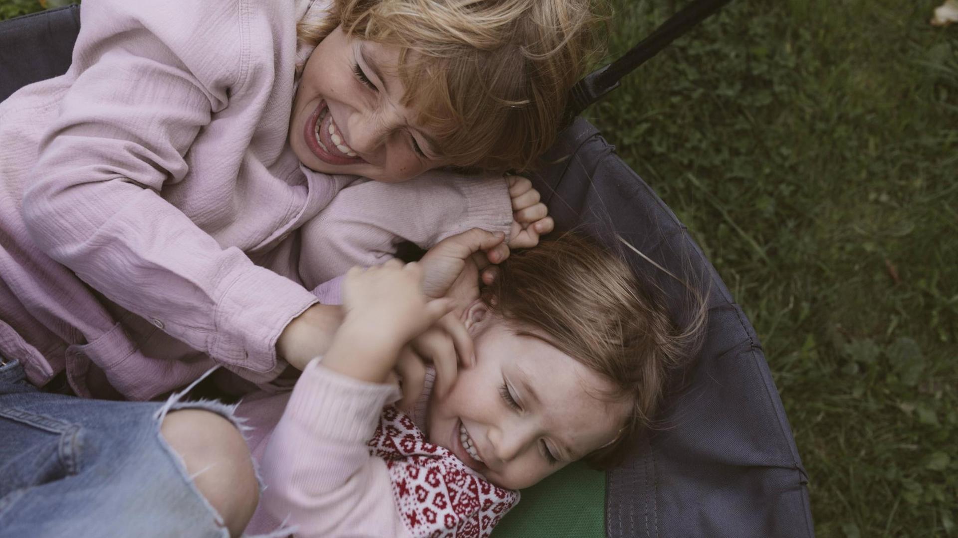  Ein kleines Mädchen und sein großer Bruder rangeln und kitzeln sich in einer Hängeschaukel. Wiese im Hintergrund.