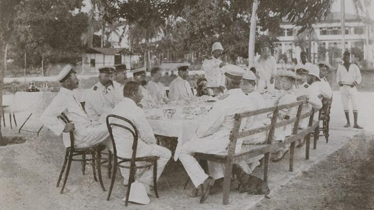 Weiß gekleidete Kolonialbeamte im damaligen Deutsch-Ostafrika sitzen um einen Tisch herum. Das Foto enstand im Jahr 1893.