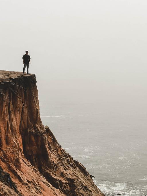Ein Mensch steht an einer Klippe und schaut aufs nebelige, graue Meer hinaus.