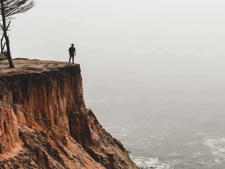 Ein Mensch steht an einer Klippe und schaut aufs nebelige, graue Meer hinaus.