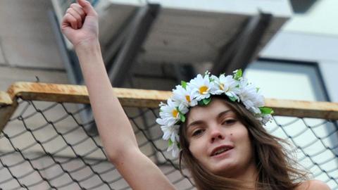 Das Schwarz-Weiß-Foto zeigt die verstorbene Femen-Aktivistin Oksana Schatschko während einer Aktion im Jahr 2011. Sie trägt einen Blumenkranz und reckt ihren rechten Arm mit geballter Faust in die Höhe.