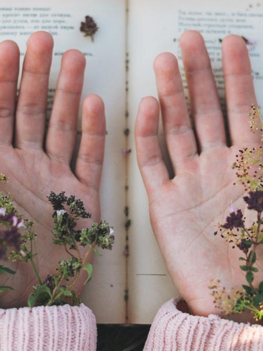 Ein Buch, darüber zwei Hände mit den Handflächen nach oben, darin liegen Blumen.