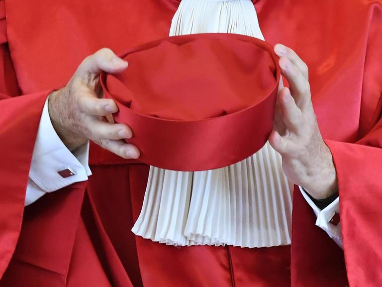 Zu sehen ist ein roter Richterhut in den Armen eines Richters in Robe.