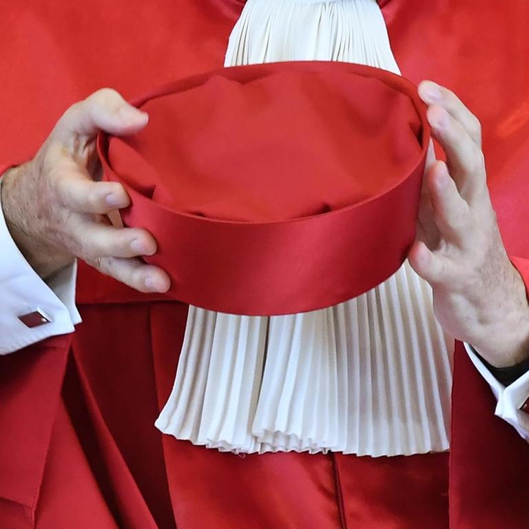 Zu sehen ist ein roter Richterhut in den Armen eines Richters in Robe.