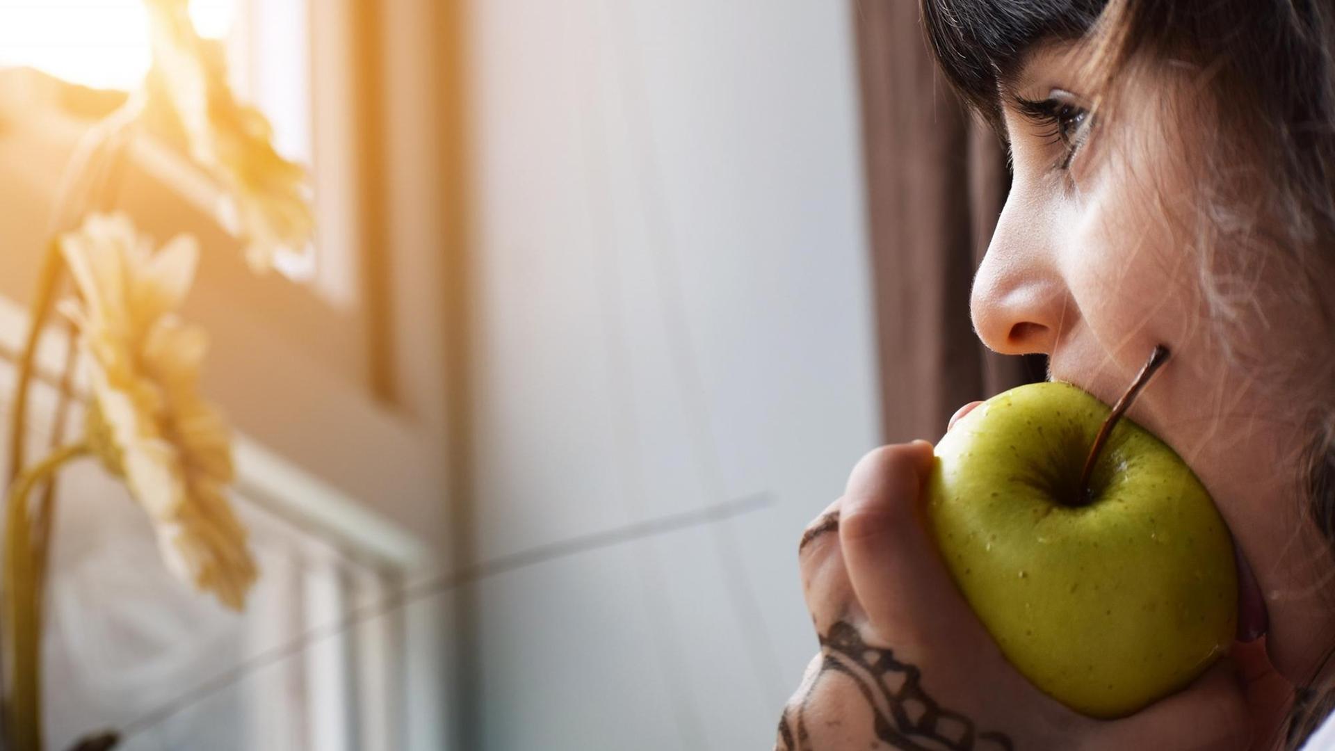 Eine junge Frau isst einen Apfel während sie am Fenster steht. (Symbolbild)