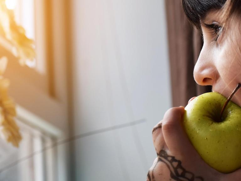 Eine junge Frau isst einen Apfel während sie am Fenster steht. (Symbolbild)