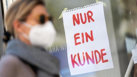 Eine Frau mit Nase-Mund-Schutzmaske geht in München an einem Geschäft vorbei, an dem ein Schild mit der Aufschrift "Nur ein Kunde" angebracht ist.