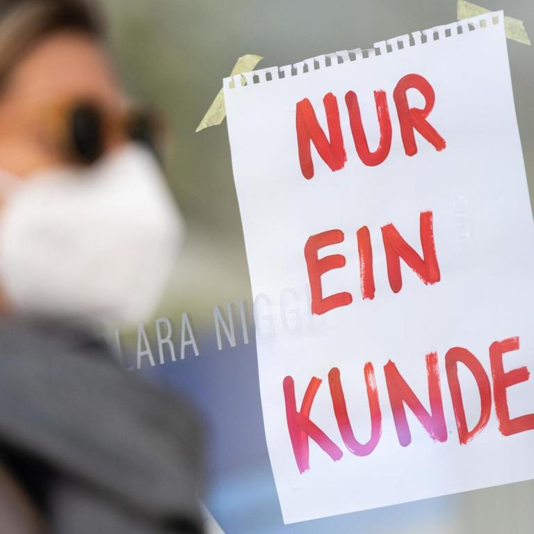 Eine Frau mit Nase-Mund-Schutzmaske geht in München an einem Geschäft vorbei, an dem ein Schild mit der Aufschrift "Nur ein Kunde" angebracht ist.