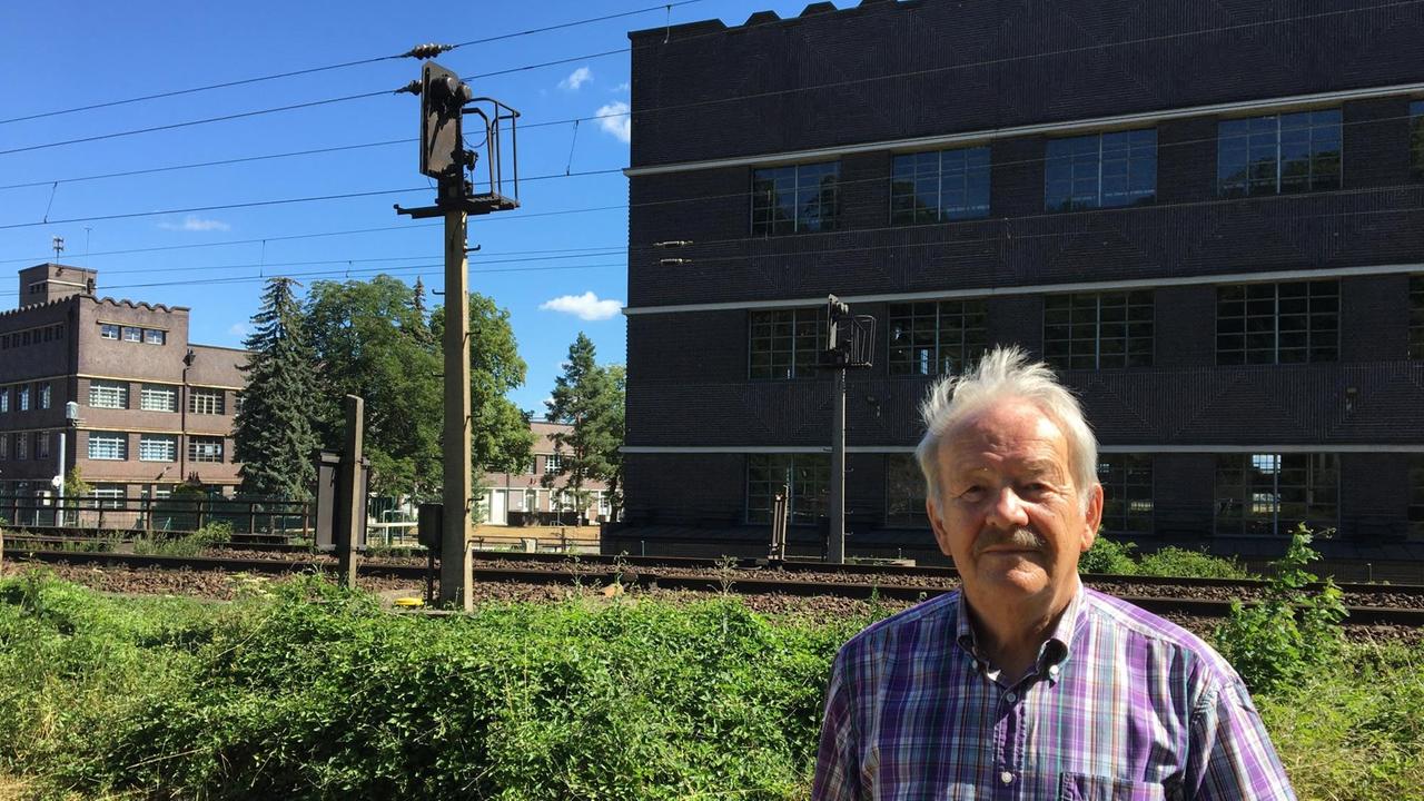 Ein älterer Mann in einem karierten Kurzarmhemd steht vor einem Bahngleis. Im Hintergrund dunkel geklinkerte Industriebauten.