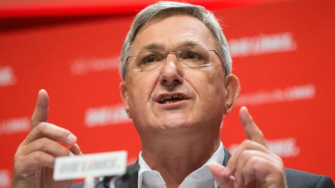 Der Vorsitzende der Partei Die Linke, Bernd Riexinger, steht vor dem Parteilogo am Rednerpult und hebt beide Zeigefinger.