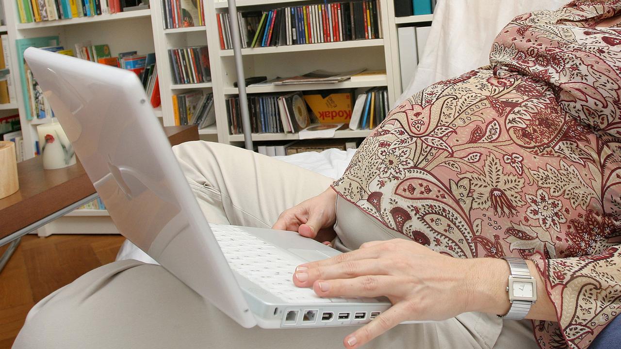 Eine schwangere Frau sitzt mit einem Laptop auf dem Sofa. 