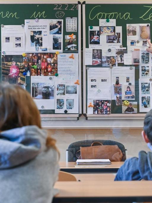 Unterrichtsszene: In einem Klassenzimmer sieht man zwei Kinder von hinten. Sie sitzen jeweils einzeln an einer Bank und blicken nach vorne zur Tafel, neben der ihre Lehrerin steht.